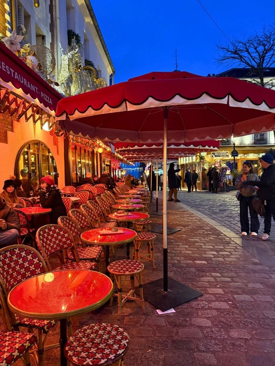 Charming Paris caf&eacute; street at dusk with red umbrellas, glowing bistro tables, and festive winter lights along cobblestone pavement.