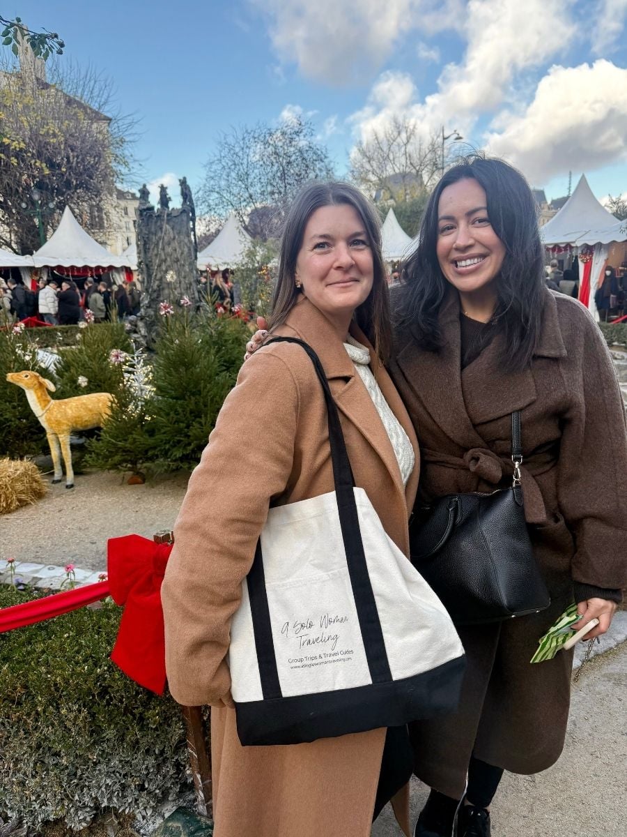 Two women smiling at a Paris holiday market with festive stalls, winter greenery, and seasonal decorations in the background.
