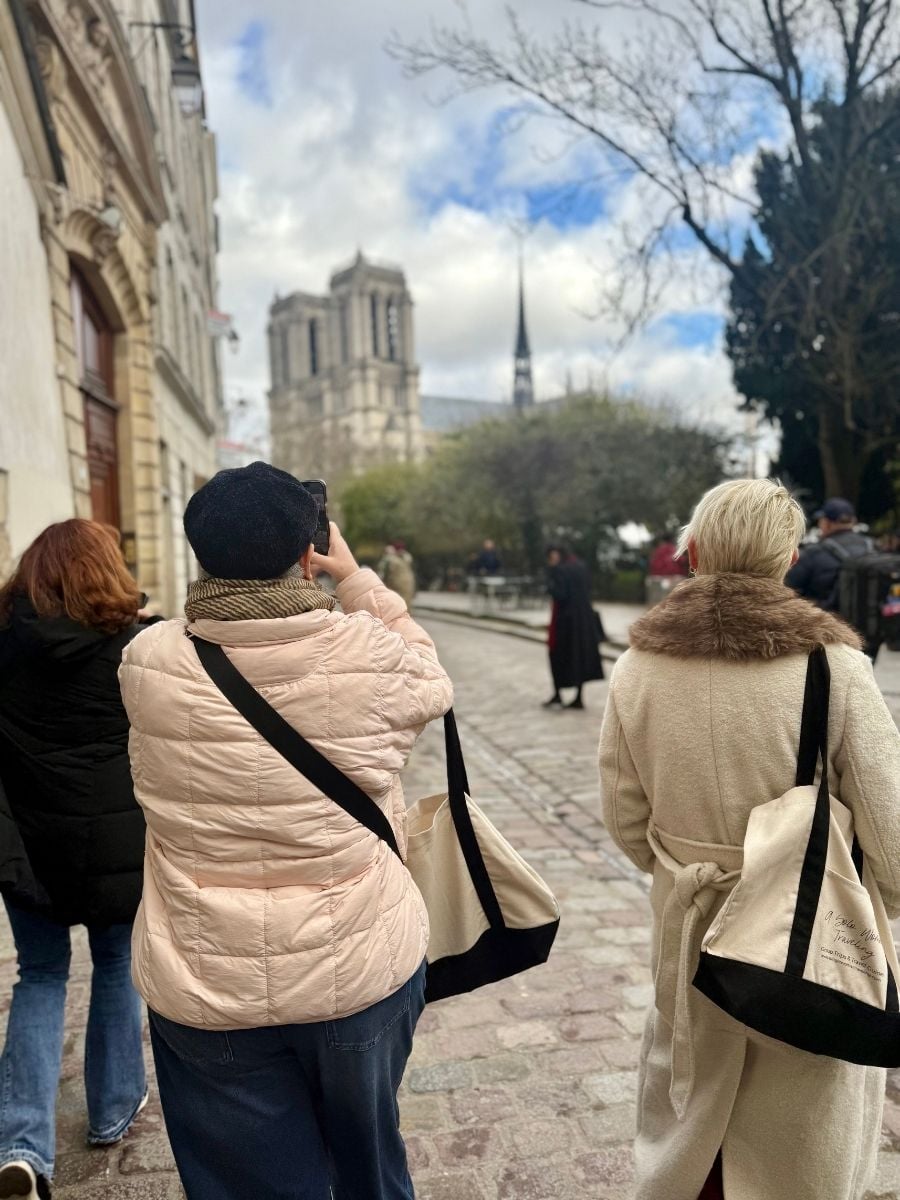 Travelers walking near Notre Dame Cathedral in Paris, carrying tote bags, with the cathedral towers visible in the distance.