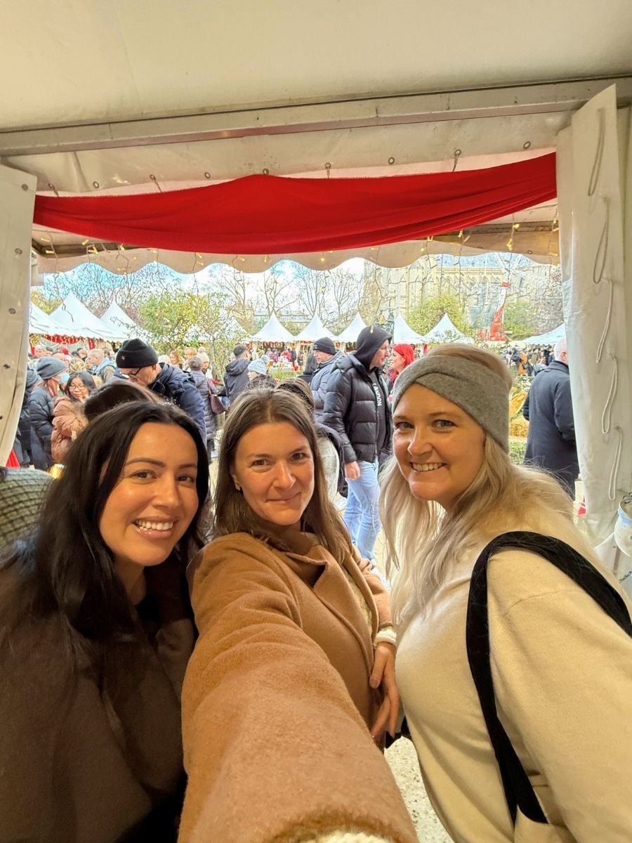 Three women taking a selfie inside a Christmas market stall in Paris with holiday tents and crowds in the background.
