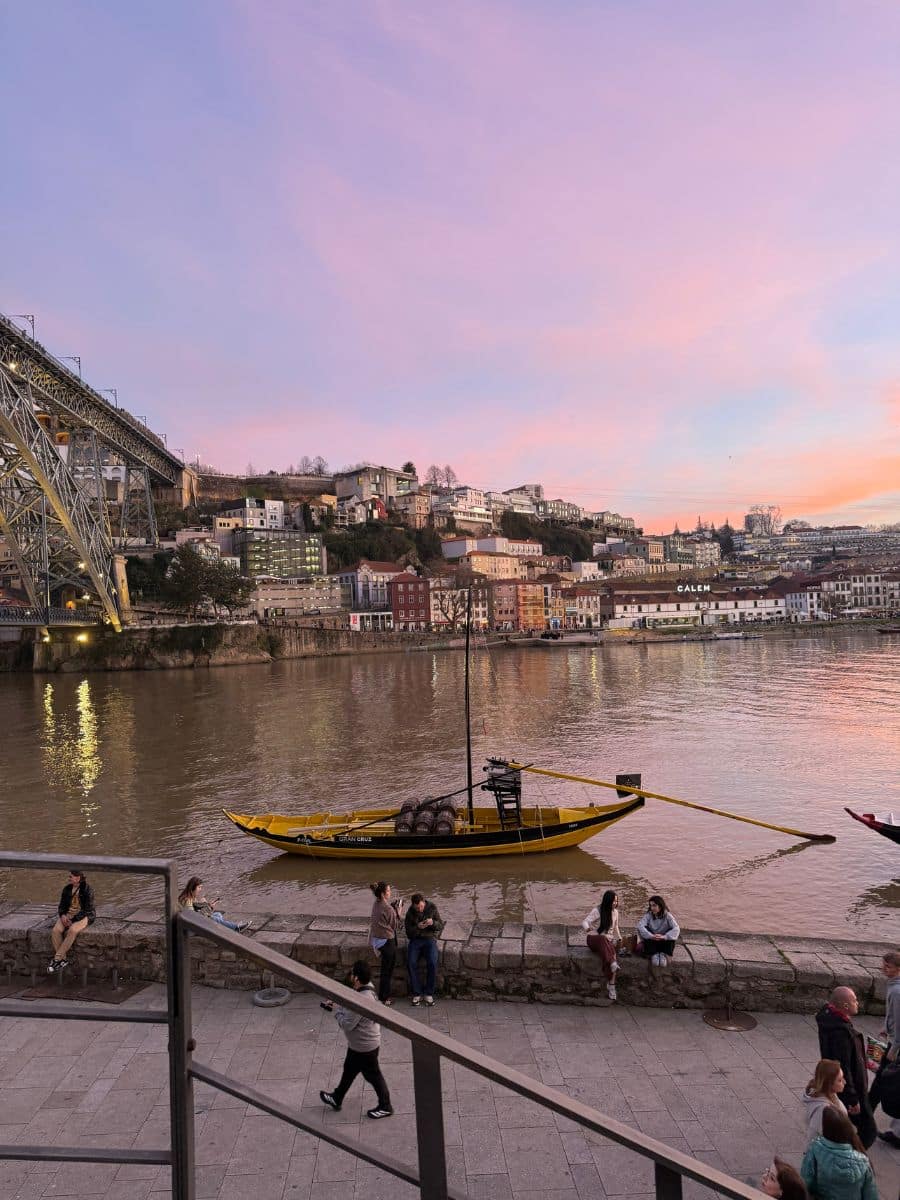 Sunset over the Douro River in Porto, with a traditional rabelo boat floating in the foreground, people relaxing along the riverfront, and the Dom Lu&iacute;s I Bridge and colorful hillside buildings glowing under a pink and purple sky.