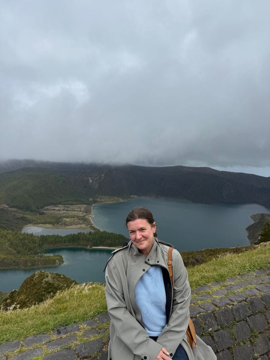 Melissa sitting at a scenic viewpoint overlooking Sete Cidades lakes in S&atilde;o Miguel, Azores with misty mountains and lush greenery