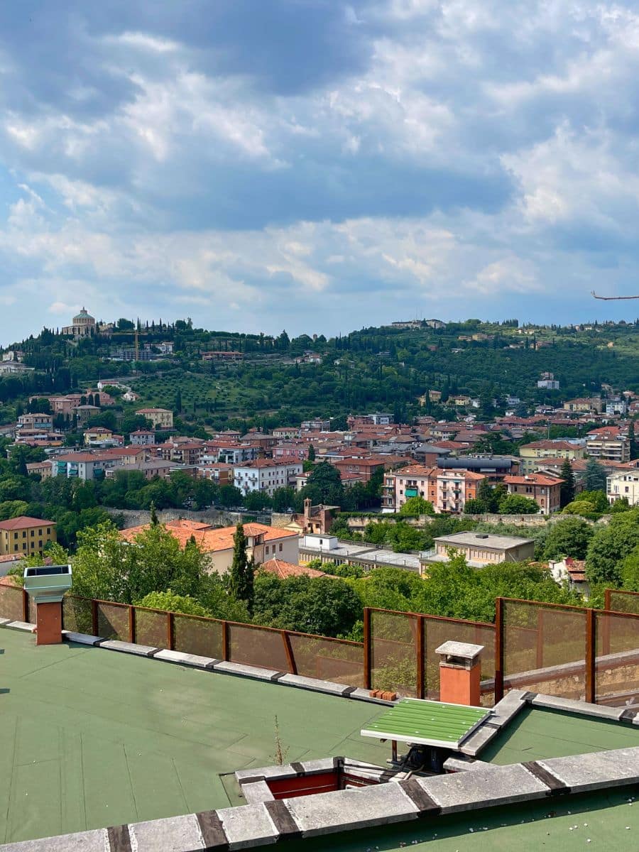 View over Verona&rsquo;s hillside neighborhoods with terracotta rooftops, green landscape, and cloudy sky