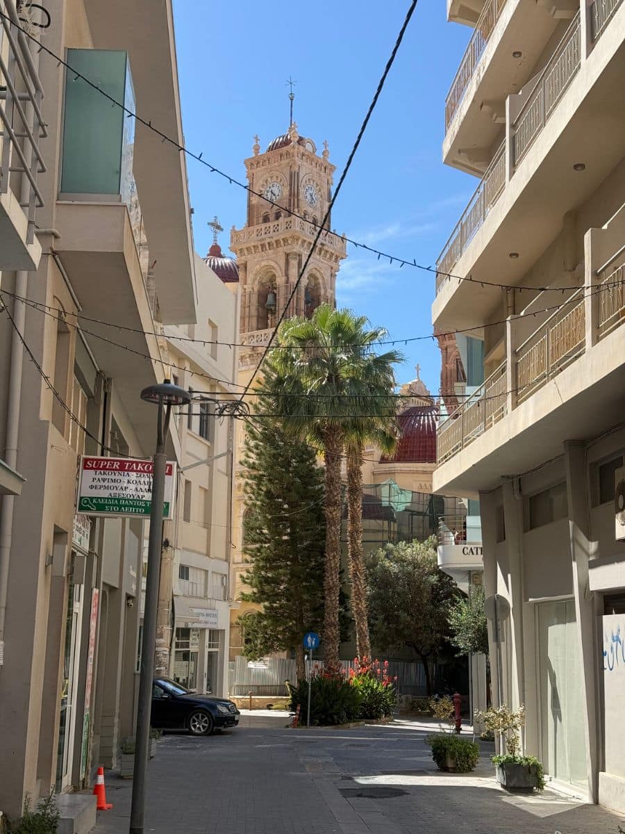 Street view of the Cathedral of Saint Minas framed by modern buildings and palm trees in Heraklion, Crete.