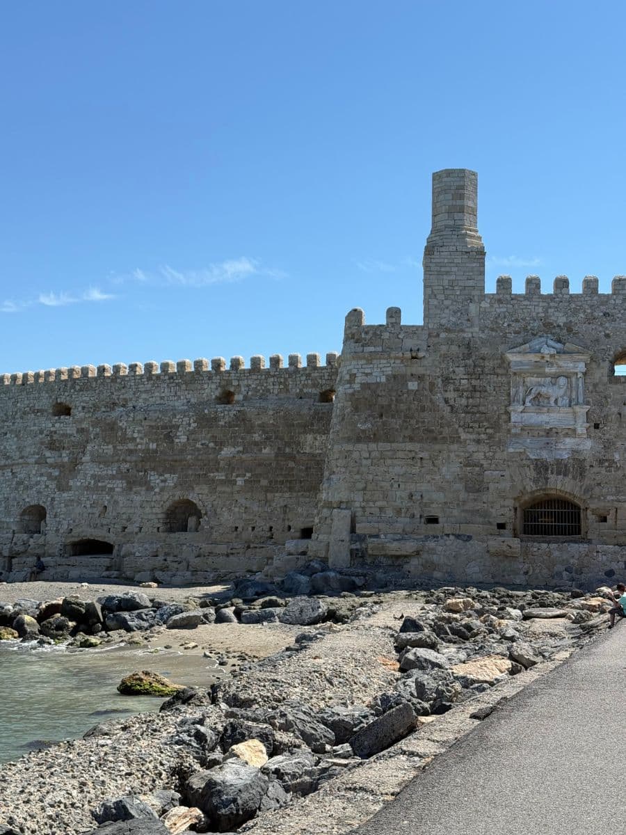 The historic Koules Fortress in Heraklion, Crete, standing along the rocky shoreline with stone walls overlooking the Mediterranean Sea under a clear blue sky.
