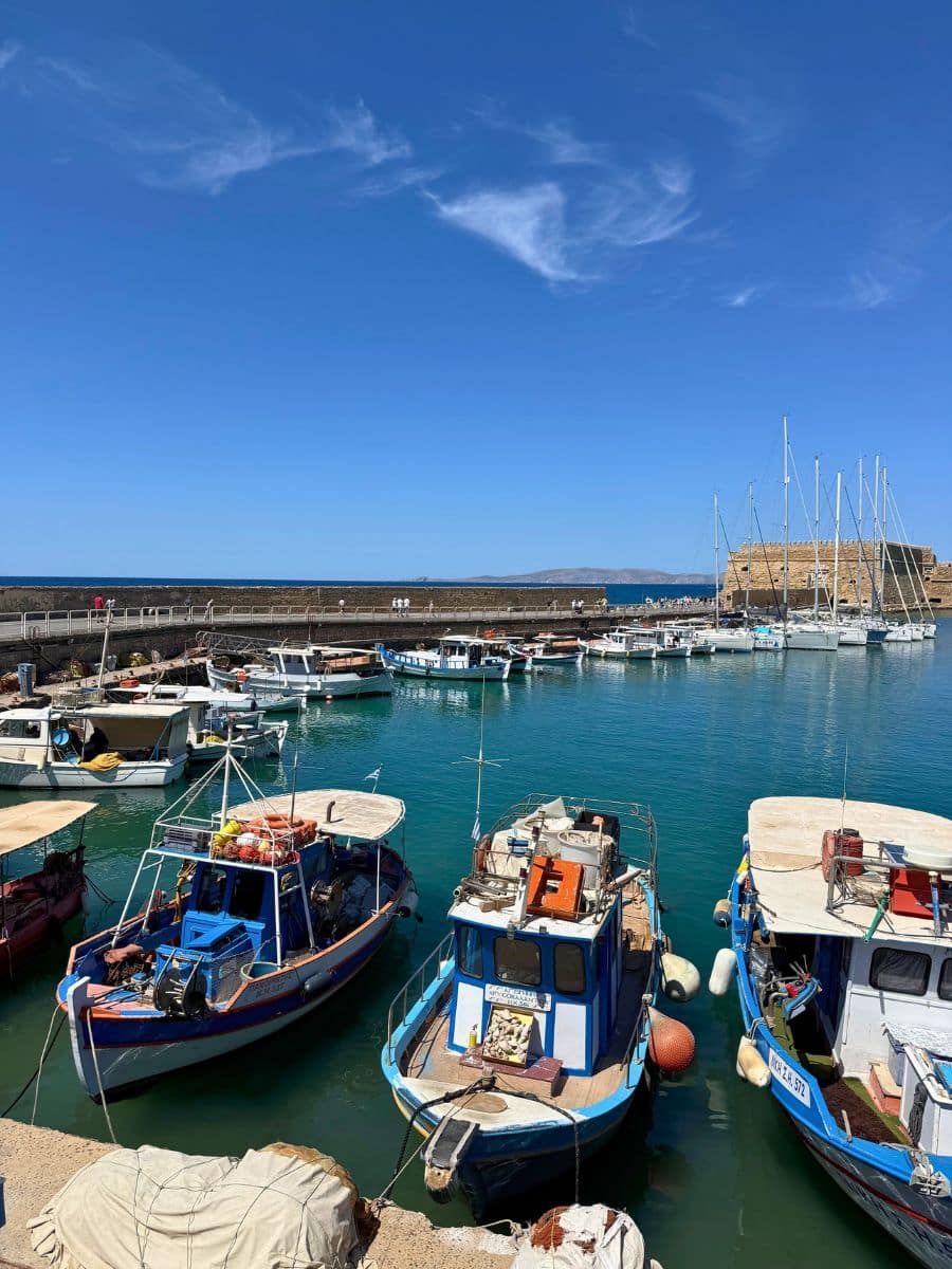 Colorful fishing boats and sailboats moored in Heraklion's Venetian Harbor, Crete, with turquoise water, the harbor walls, and Koules Fortress in the background.