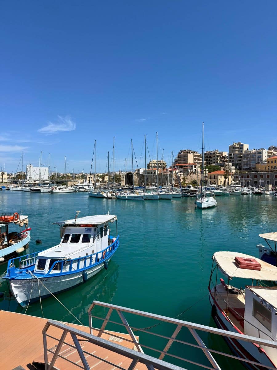 Panoramic view of Heraklion's Venetian Harbor in Crete with fishing boats, sailboats, calm turquoise water, and the city skyline under a bright blue sky.