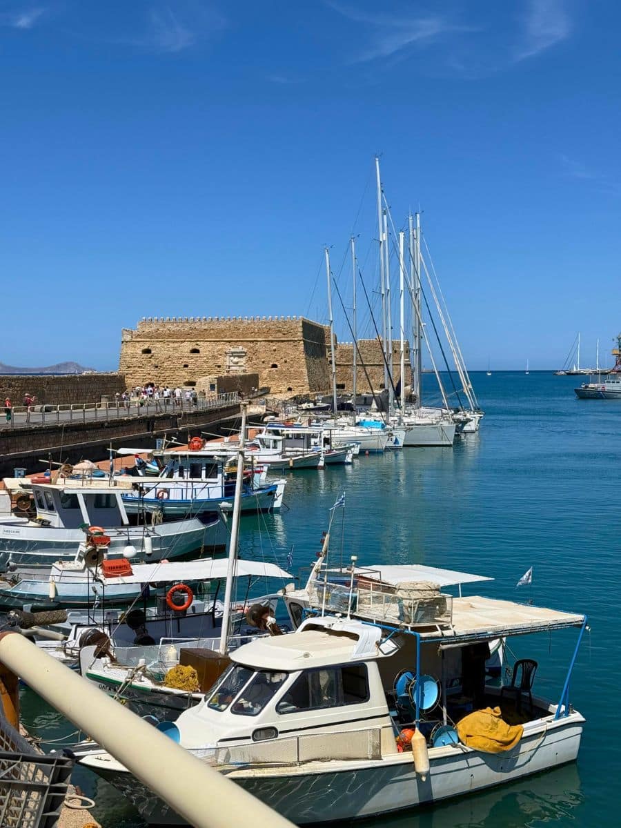 Fishing boats and sailboats docked beside Koules Fortress in Heraklion's Venetian Harbor, Crete, with the sparkling Mediterranean Sea and historic harbor walls in the background.