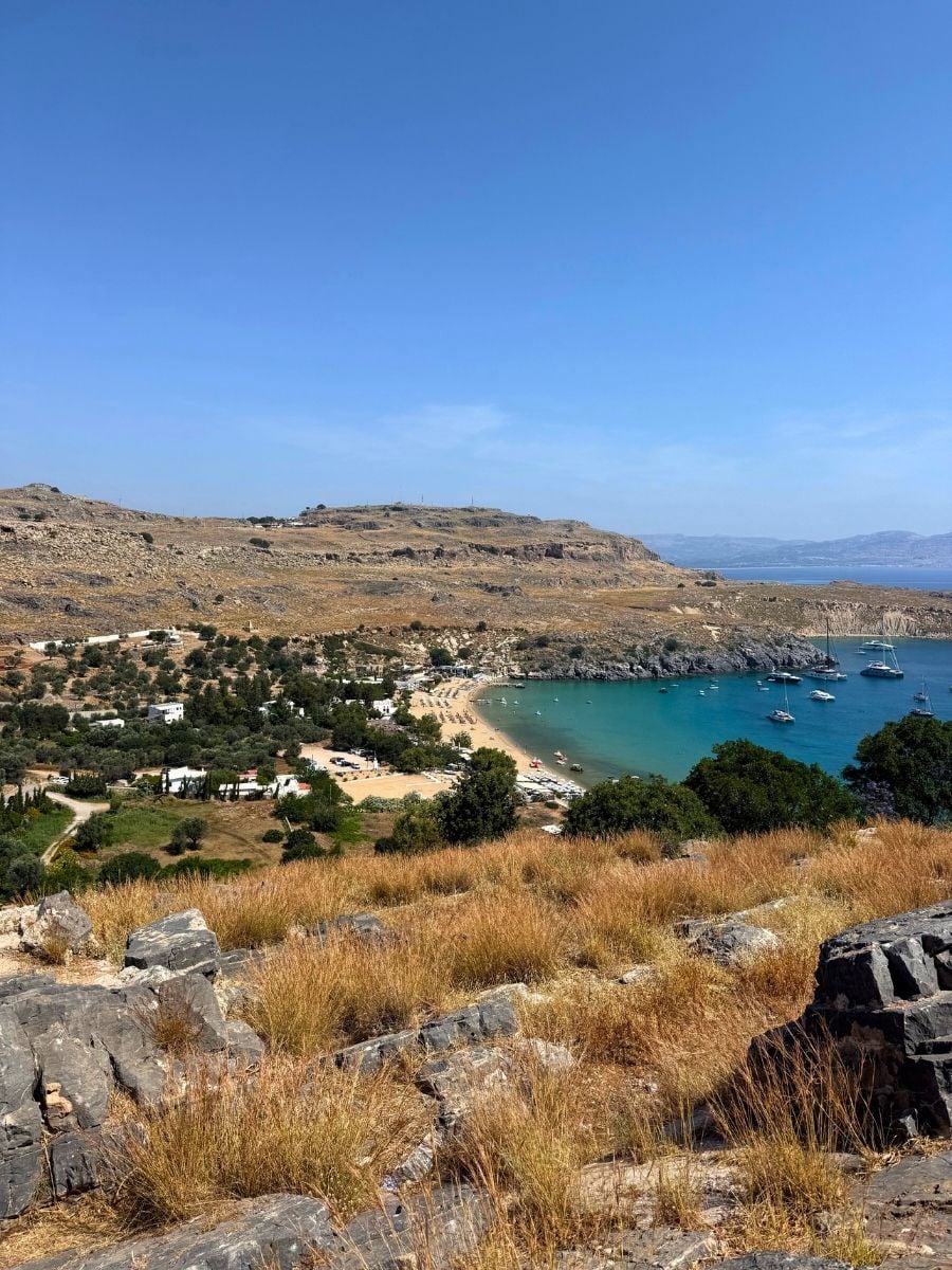 Elevated view of a sandy bay near Lindos, Rhodes, Greece with turquoise water, beach umbrellas, and anchored yachts.