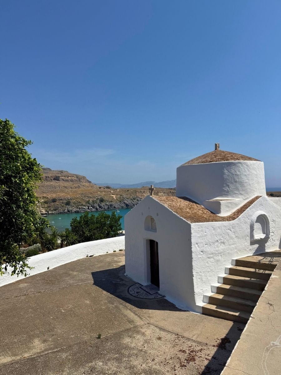 Whitewashed chapel overlooking St. Paul's Bay in Lindos, Rhodes, Greece with sweeping coastal views and calm blue water.