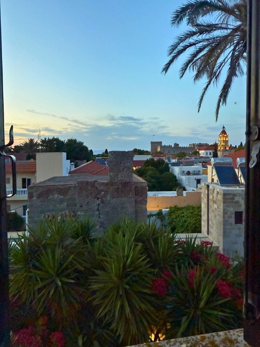 Evening view over rooftops, palm trees, and medieval walls from Rhodes Old Town, Rhodes, Greece at dusk.