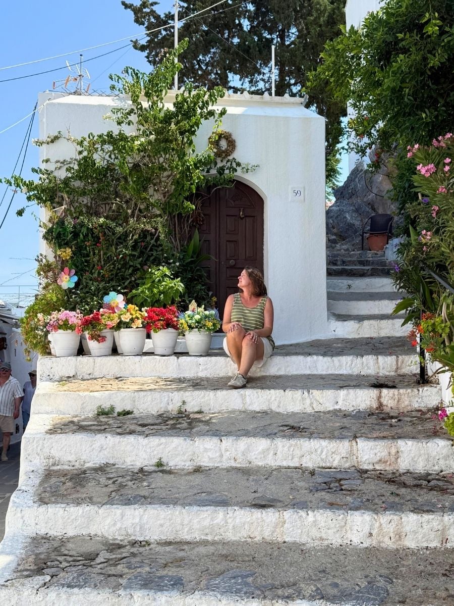 Whitewashed home with colorful flower pots in Lindos, Rhodes, Greece where a visitor relaxes on sunlit stone steps.