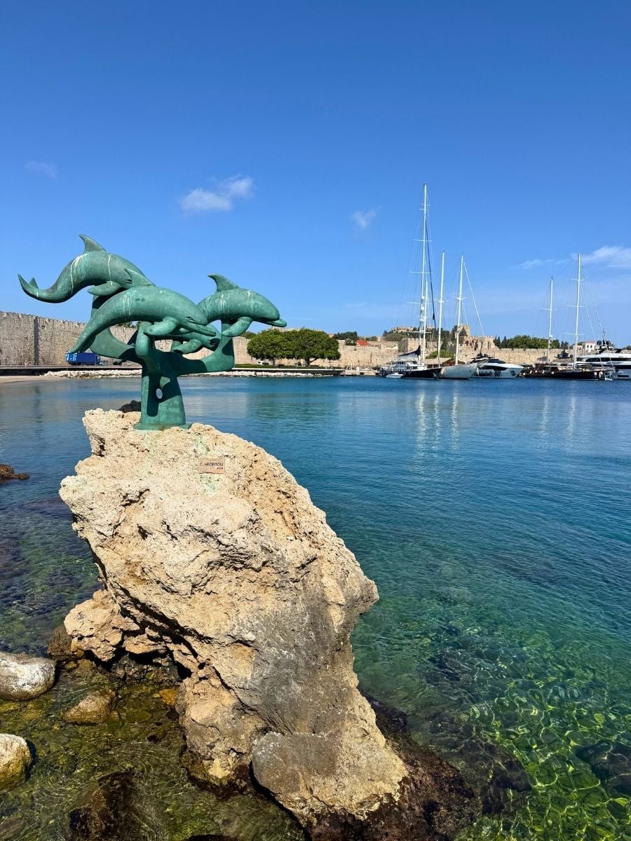Bronze dolphin sculpture overlooking crystal-clear water and the harbor in Rhodes Town, Rhodes, Greece.