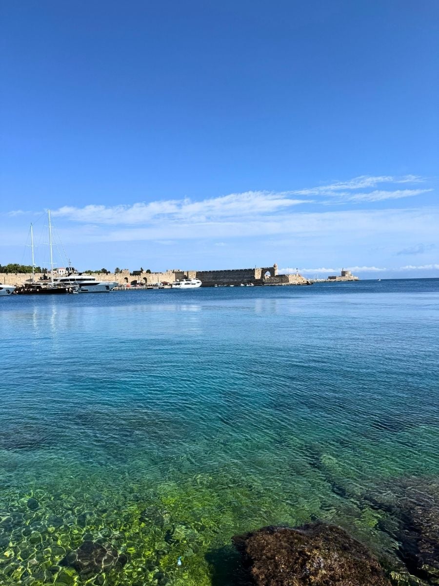 Clear turquoise waters and the fortified harbor walls of Rhodes Town, Rhodes, Greece under a cloudless sky.