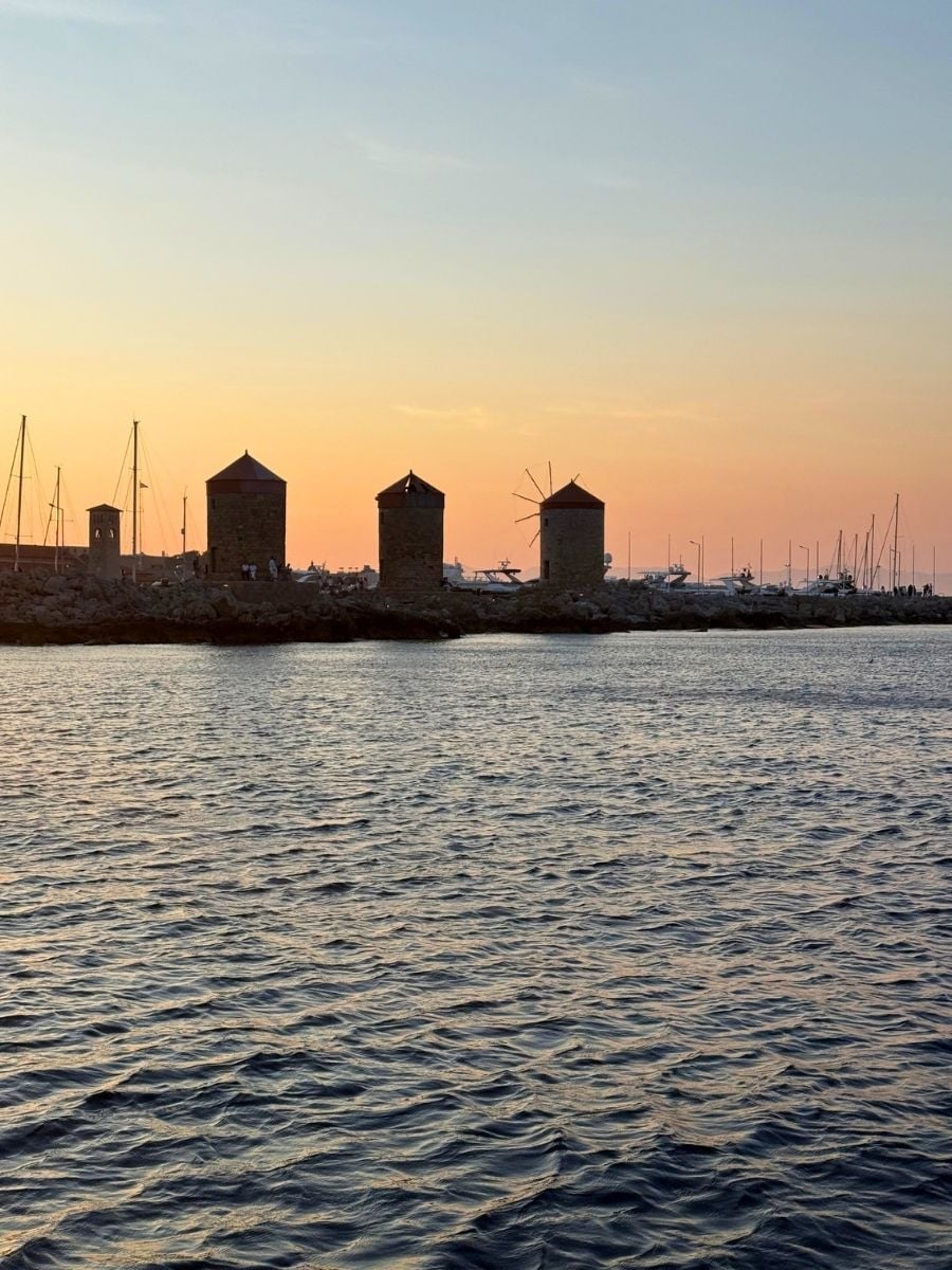 Historic windmills lining Mandraki Harbour in Rhodes, Greece, glowing in warm sunset light beside the Aegean Sea.