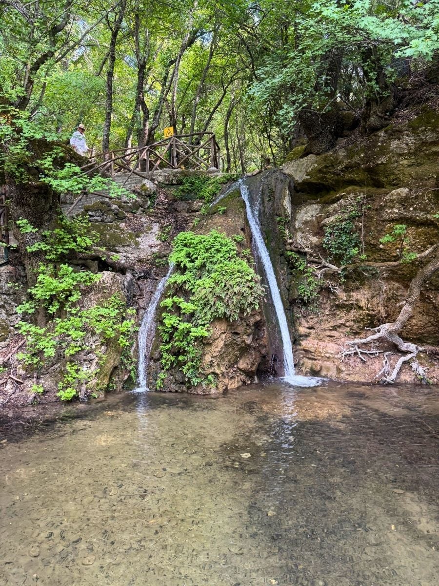 Small waterfall cascading into a clear natural pool in Seven Springs, Rhodes, Greece, surrounded by mossy rocks and woodland.
