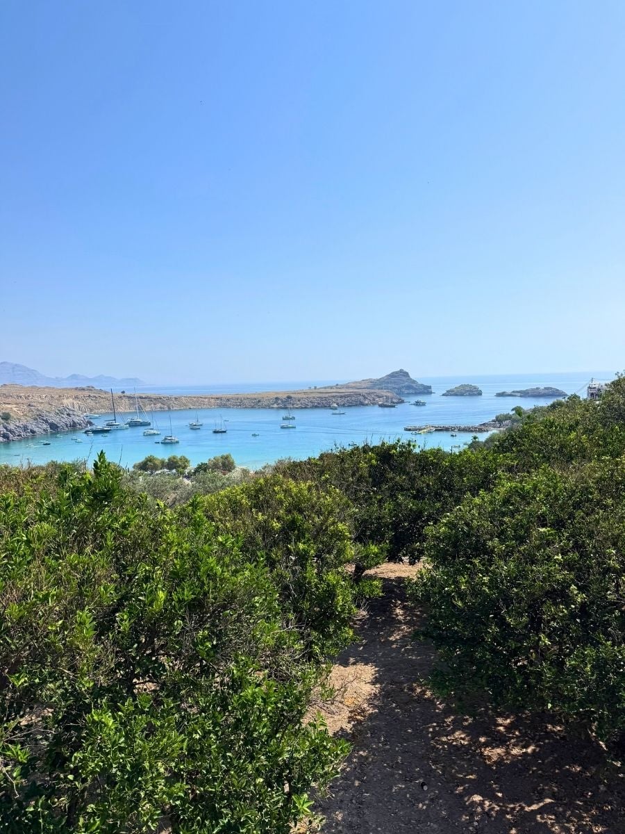 Coastal trail overlooking St. Paul's Bay in Lindos, Rhodes, Greece with turquoise water, anchored boats, and Mediterranean vegetation.