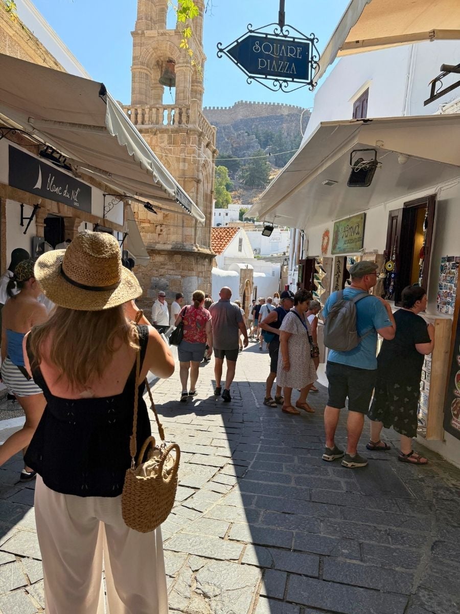 Visitors strolling through a lively pedestrian street in Lindos, Rhodes, Greece beneath the medieval acropolis and historic bell tower.