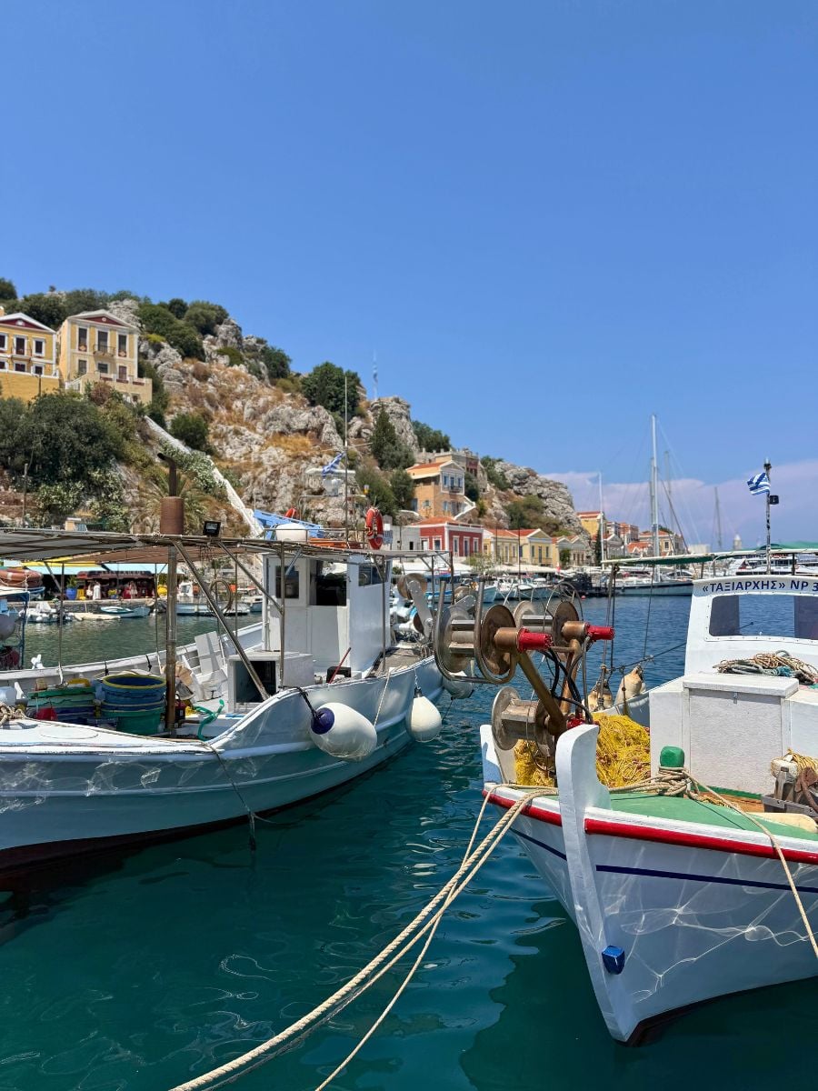 Traditional fishing boats moored in the colorful harbor of Symi, Greece, with pastel hillside houses and crystal-clear turquoise water under a bright blue sky