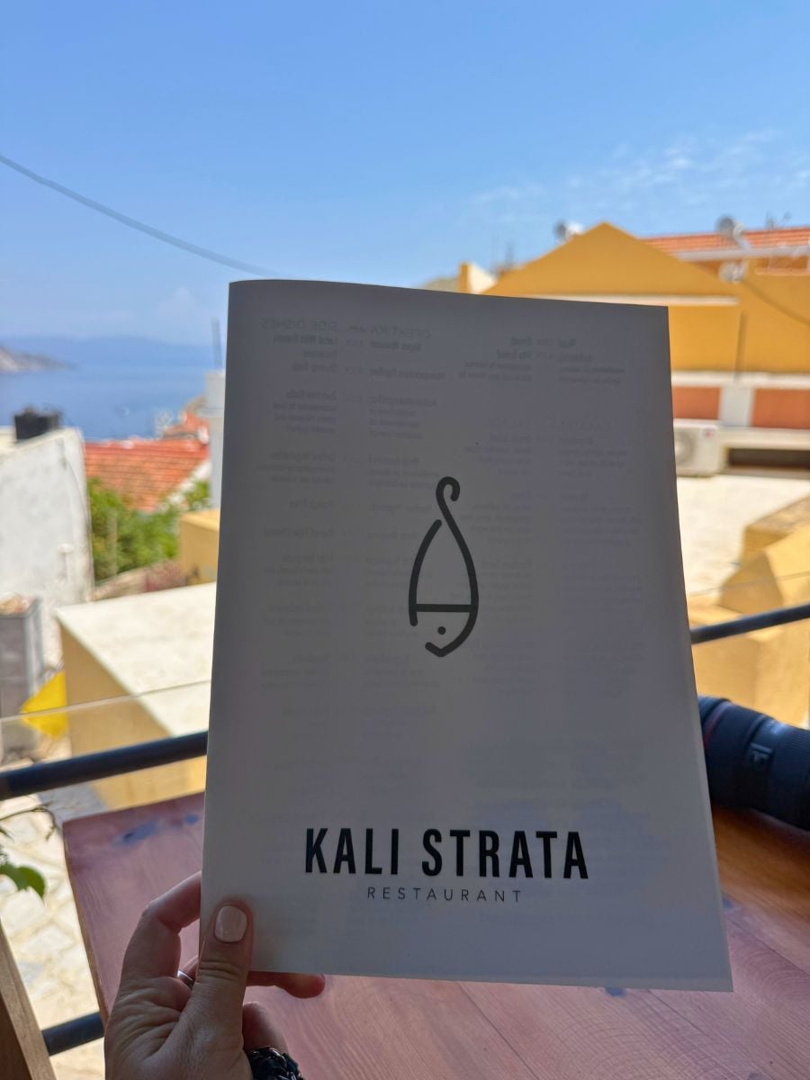 Kali Strata Restaurant menu held against scenic rooftop views of Symi, Greece and the Aegean Sea.