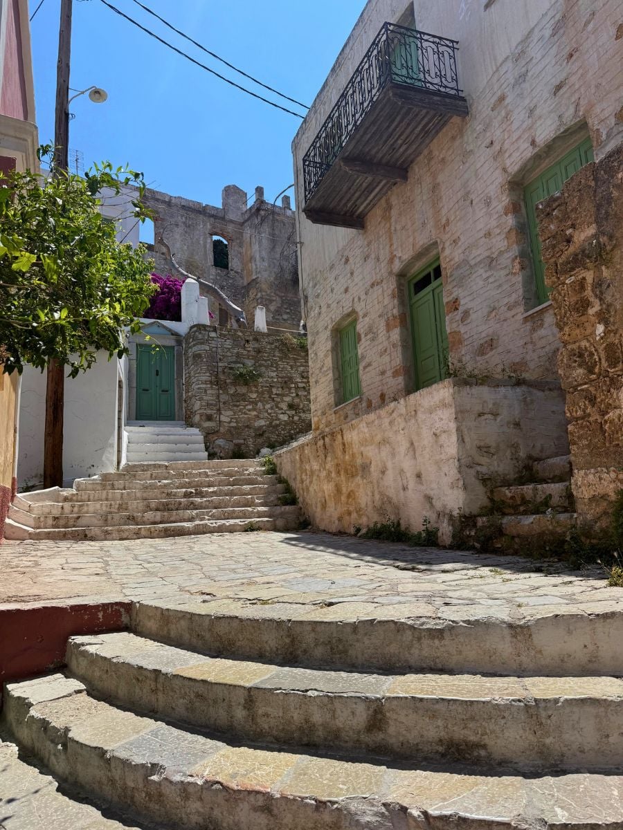 Quiet stone street in Symi, Greece lined with historic buildings, green shutters, and worn stone steps leading uphill.
