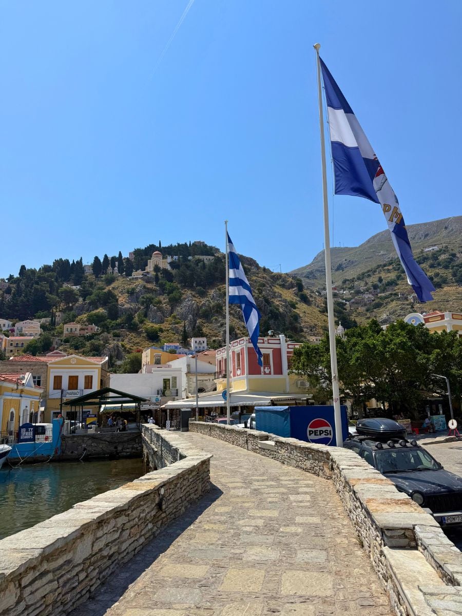 Greek flags waving above the waterfront promenade in Symi, Greece with colorful harbor buildings and hillside churches in the background.