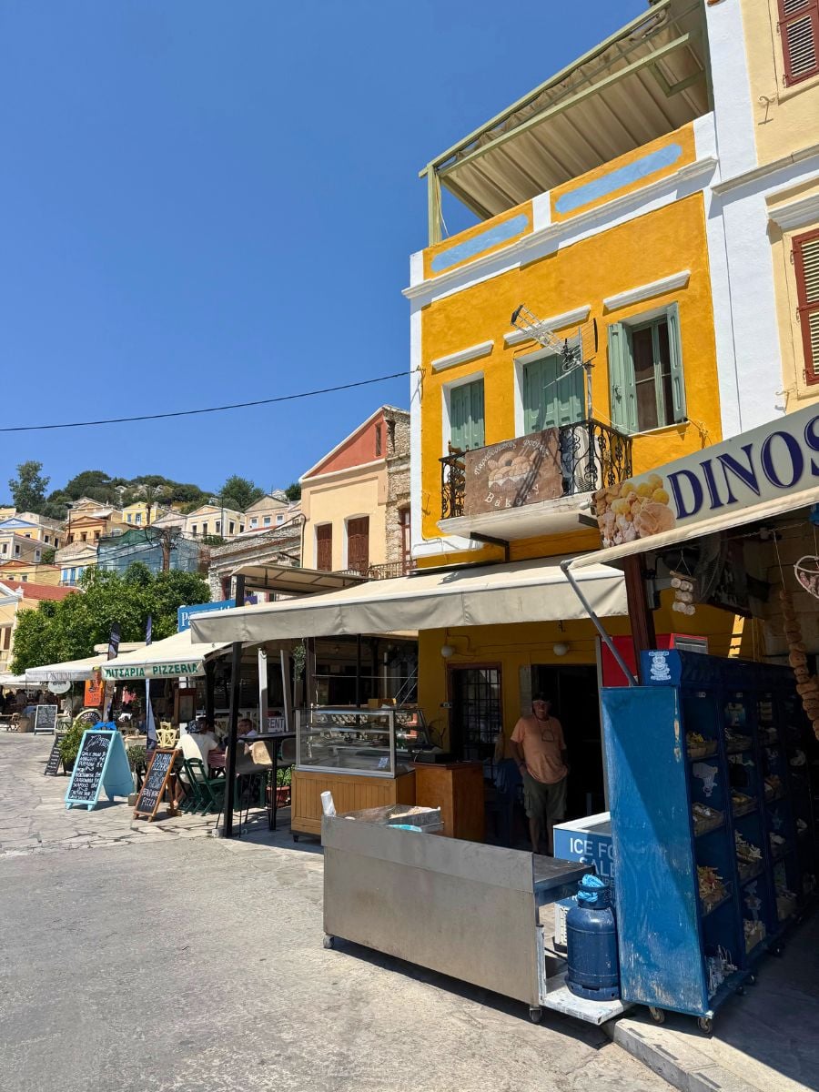 Colorful waterfront street in Symi, Greece featuring local cafés, bakeries, and traditional island architecture on a sunny day.