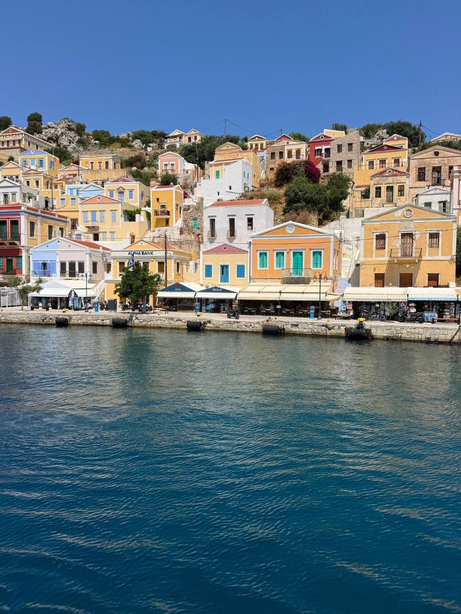 Colorful neoclassical waterfront houses in Symi, Greece reflected along the calm harbor beneath a bright blue Mediterranean sky.