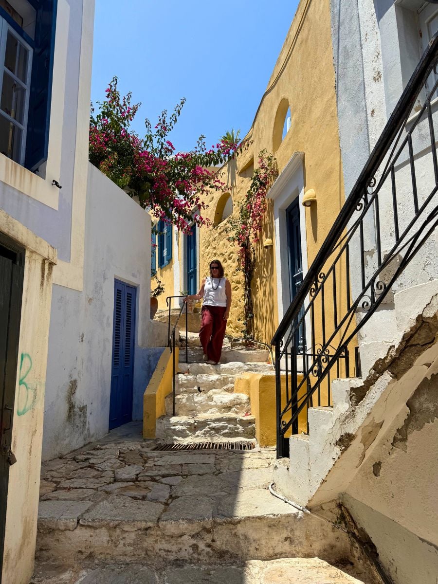 Melissa standing on charming stone staircase in Symi, Greece lined with pastel buildings, blue shutters, and blooming bougainvillea on a sunny afternoon.