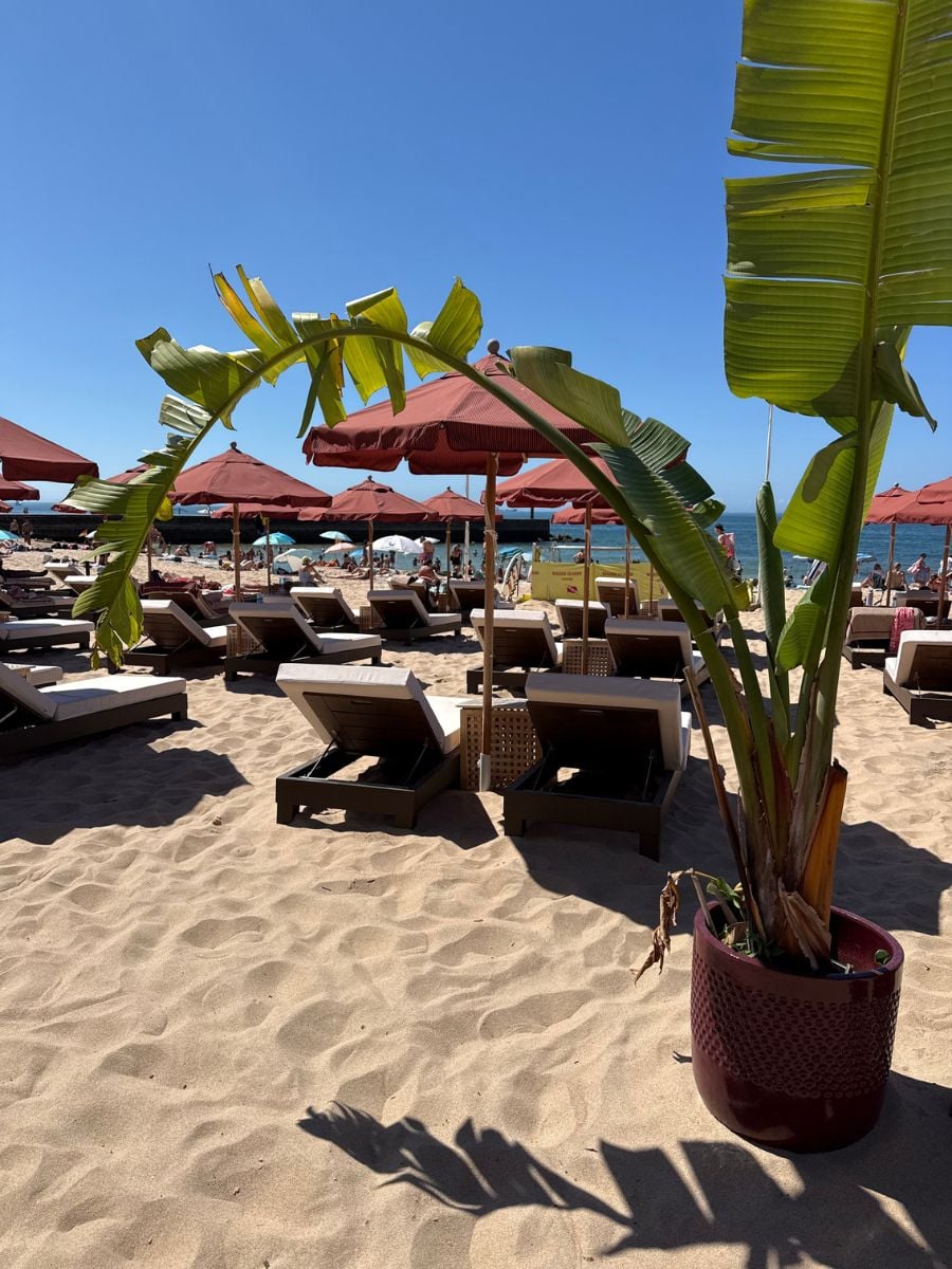 Sun loungers and red umbrellas at Época Beach Club on Cascais Beach in Estoril, Portugal under bright blue skies.